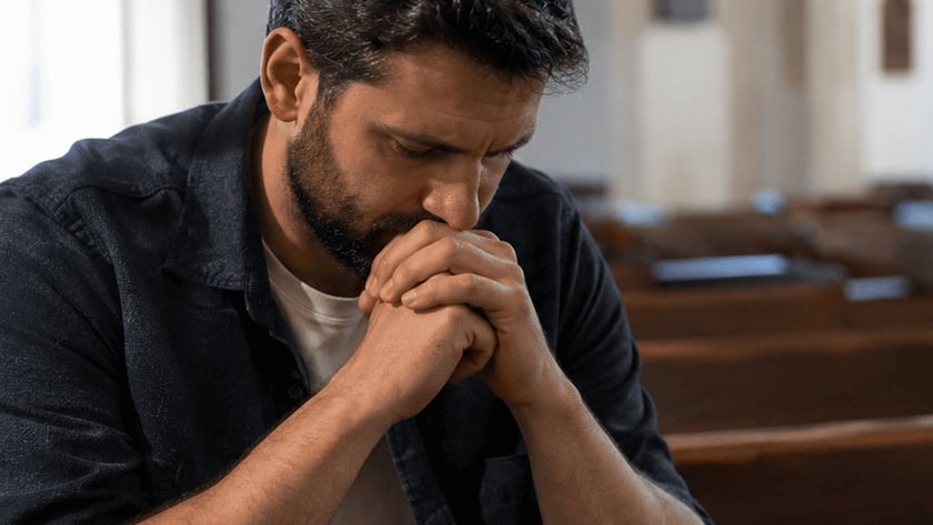 Homem de barba, com as mãos unidas junto ao rosto, sentado em um banco de igreja.