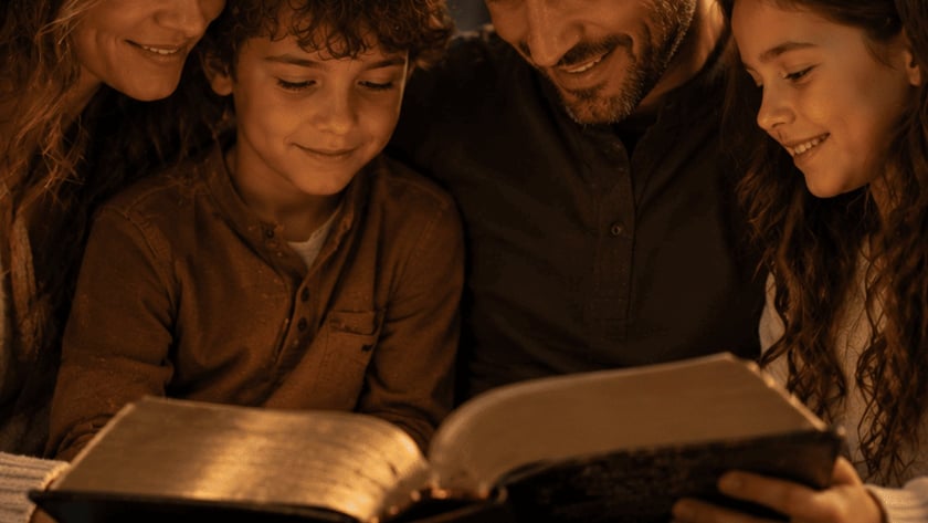 Homem sorridente, uma menina e um menino leem juntos um livro grande num sofá.