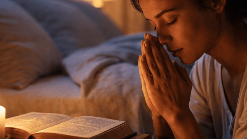 Mulher de olhos fechados e mãos juntas reza ao lado de uma Bíblia, vela e terço.