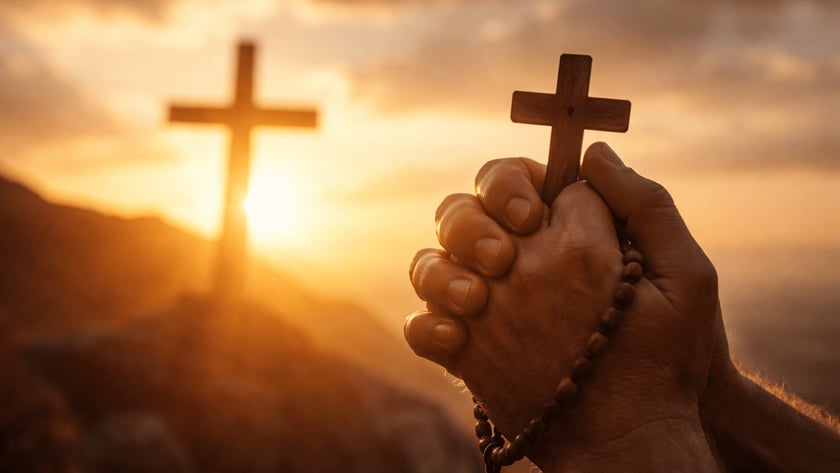 Mãos postas segurando um terço com crucifixo de madeira diante de uma cruz ao pôr do sol.