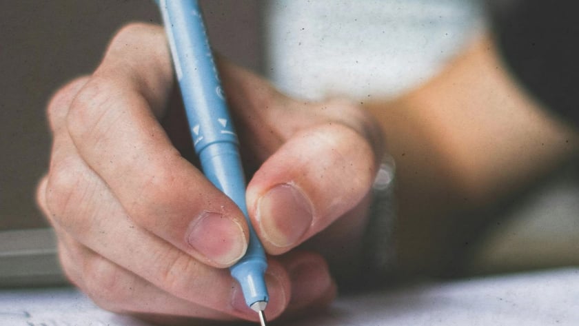 Mão segurando uma caneta azul-clara e escrevendo em um caderno aberto sobre a mesa.