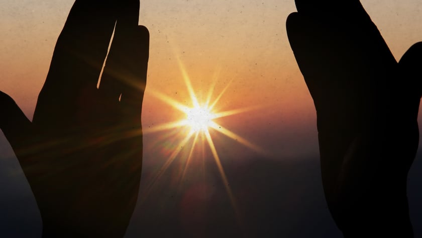 Duas mãos erguidas em silhueta enquadram o sol brilhante ao entardecer.
