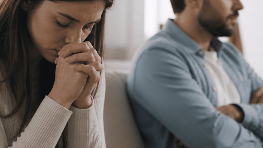 Mulher de cabeça baixa e mãos juntas, ao lado de homem de braços cruzados no sofá.