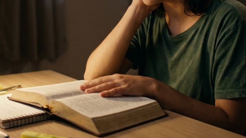 Mulher de olhos fechados, com a mão sobre um livro aberto, à mesa à noite.
