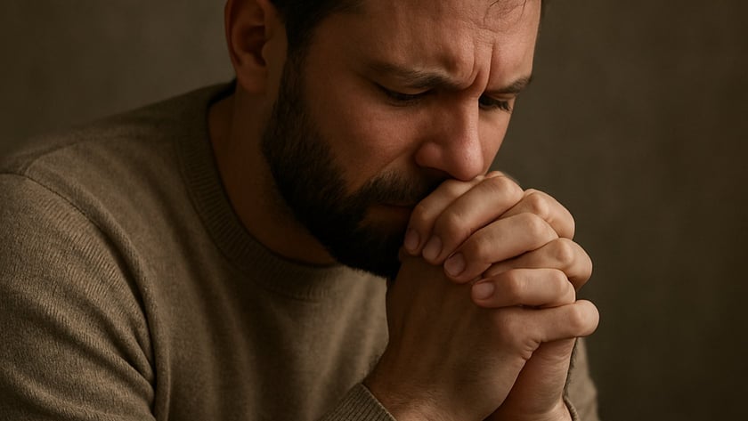 Homem de barba, com suéter marrom, de olhos fechados e mãos unidas junto ao rosto.