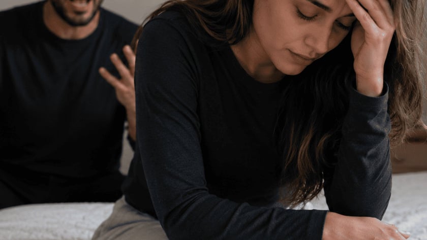 Mulher sentada na cama com a mão na testa, enquanto homem atrás grita e gesticula.