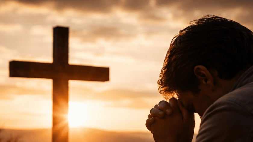 Homem de mãos postas e cabeça baixa diante de uma cruz de madeira, com velas.