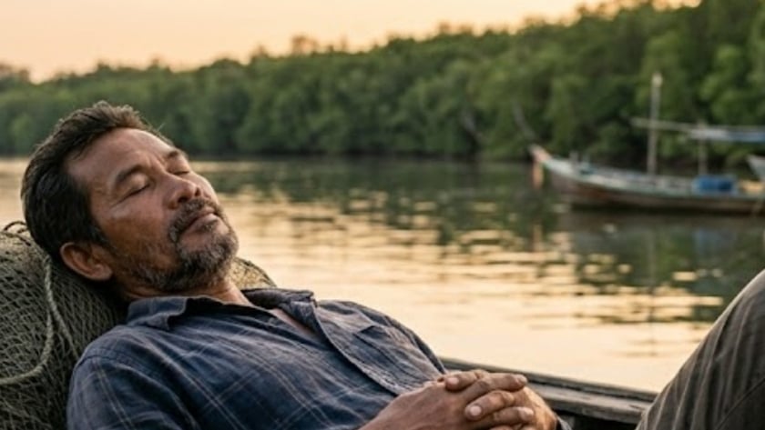 Homem de barba dorme recostado em redes de pesca num barco, perto de um rio.