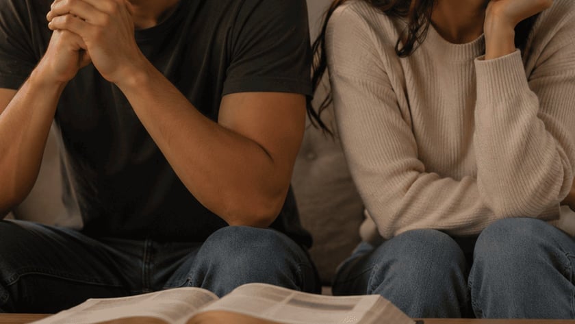 Homem e mulher sentados em um sofá, de costas um para o outro, com uma Bíblia aberta na mesa.