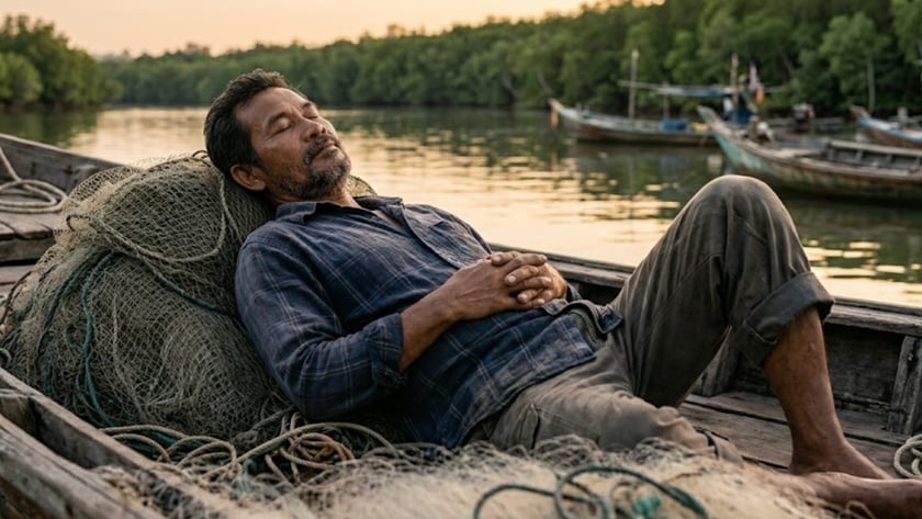 Homem de barba dorme recostado em redes de pesca num barco, perto de um rio.