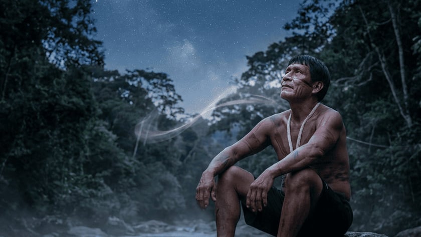 Homem indígena sentado em uma pedra à beira de um rio, olhando a lua.