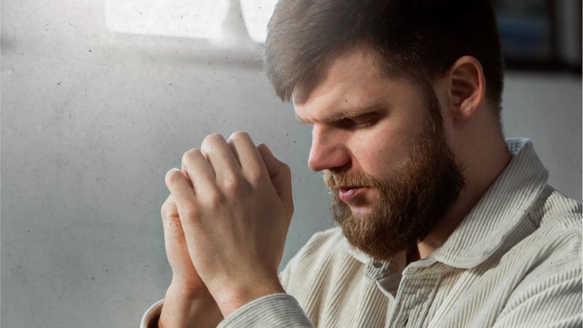 Homem barbudo de camisa bege com as mãos unidas, de olhos fechados, diante de uma Bíblia.