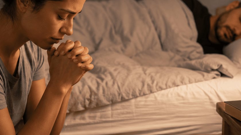Homem dorme na cama enquanto uma pessoa, em primeiro plano, ora de mãos postas.