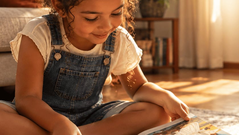 Menina de cabelo cacheado sentada no chão folheia um livro ilustrado aberto em uma sala.