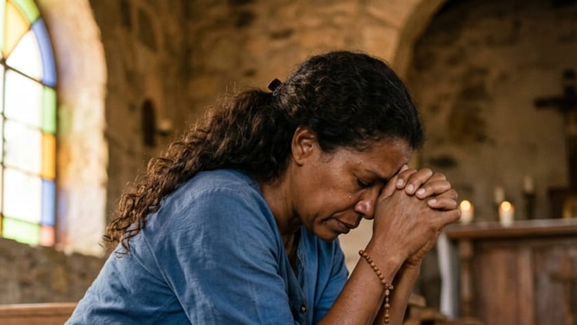 Mulher ajoelhada em uma igreja de pedra, com as mãos postas e um terço no pulso.
