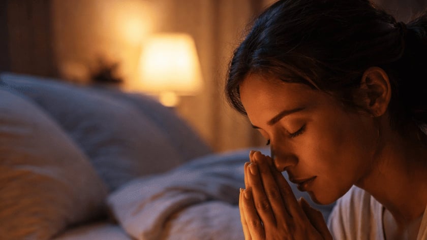 Mulher de olhos fechados e mãos juntas reza ao lado de uma Bíblia, vela e terço.