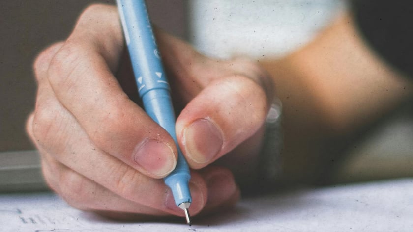 Mão segurando uma caneta azul-clara e escrevendo em um caderno aberto sobre a mesa.