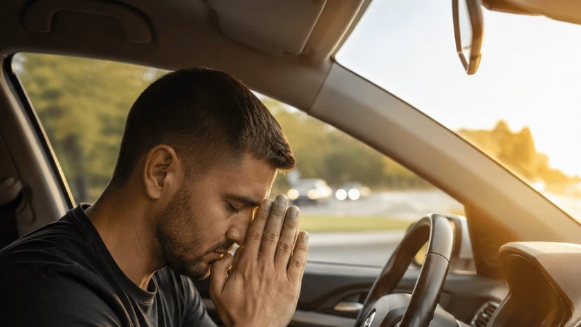 Homem de camiseta escura sentado ao volante, com as mãos unidas diante do rosto e olhos fechados.