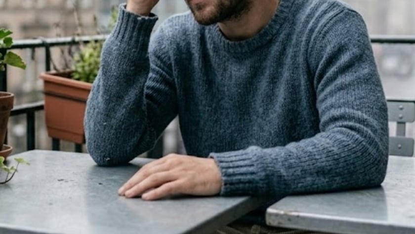 Homem barbado de suéter azul sentado a uma mesa de metal, com a mão na testa.