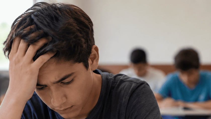 Estudante de cabelo escuro, com a mão na testa, olha para um caderno em sala de aula.