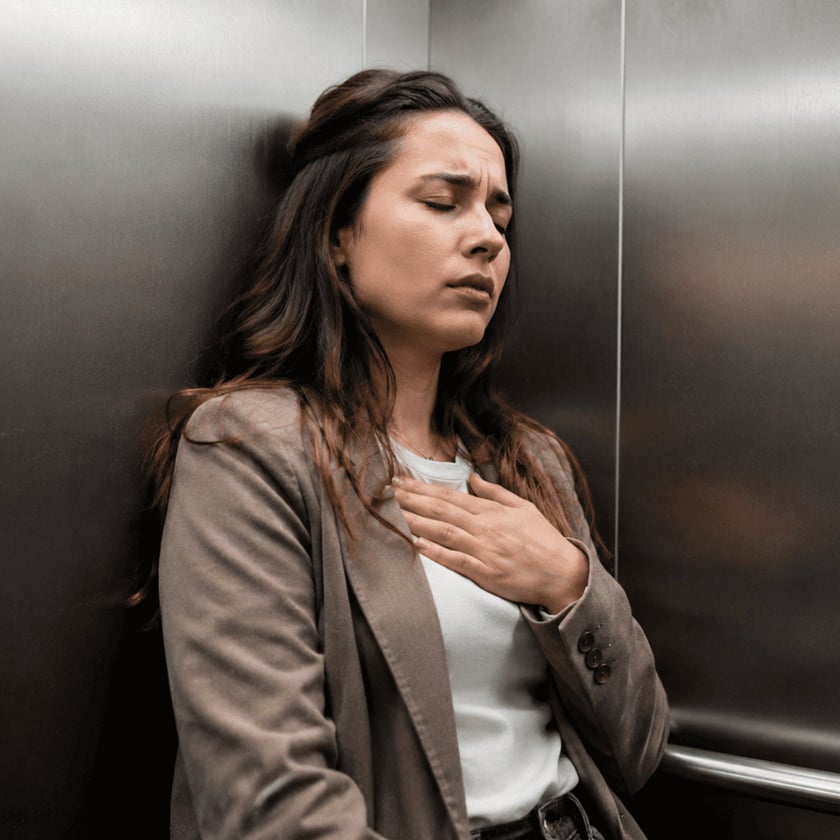 Mulher de blazer marrom encostada na parede de um elevador, com a mão no peito.