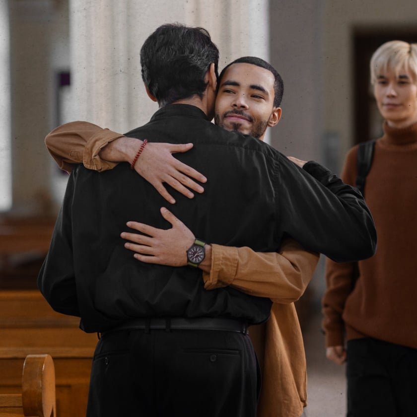 Dois homens se abraçam em uma igreja, enquanto um jovem loiro os observa ao fundo.