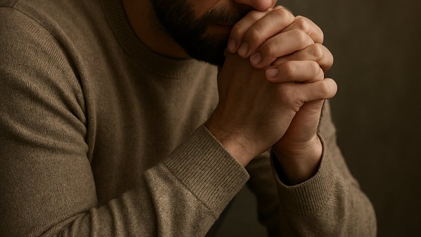 Homem de barba, com suéter marrom, de olhos fechados e mãos unidas junto ao rosto.