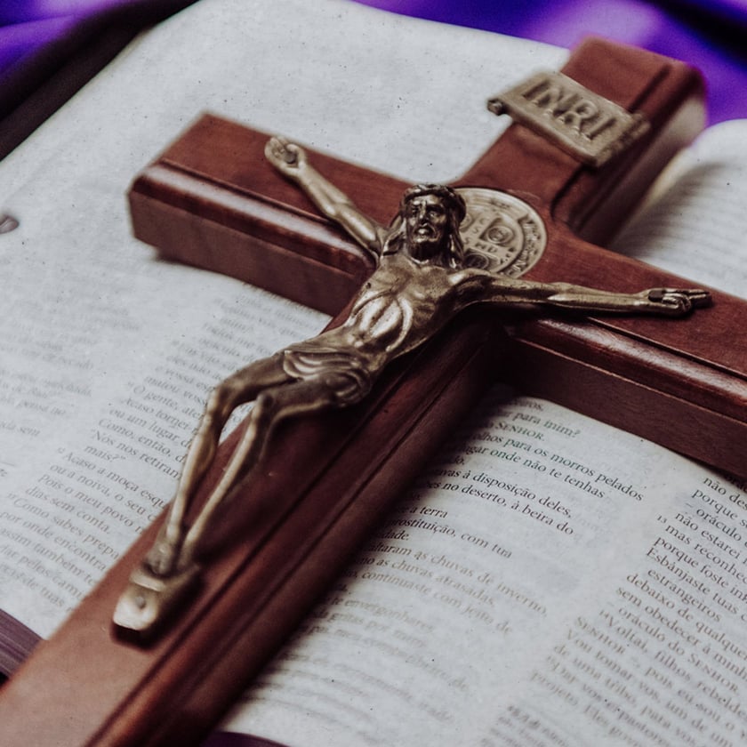 Crucifixo de madeira com Cristo metálico sobre Bíblia aberta, com tecido roxo ao fundo. "INRI"