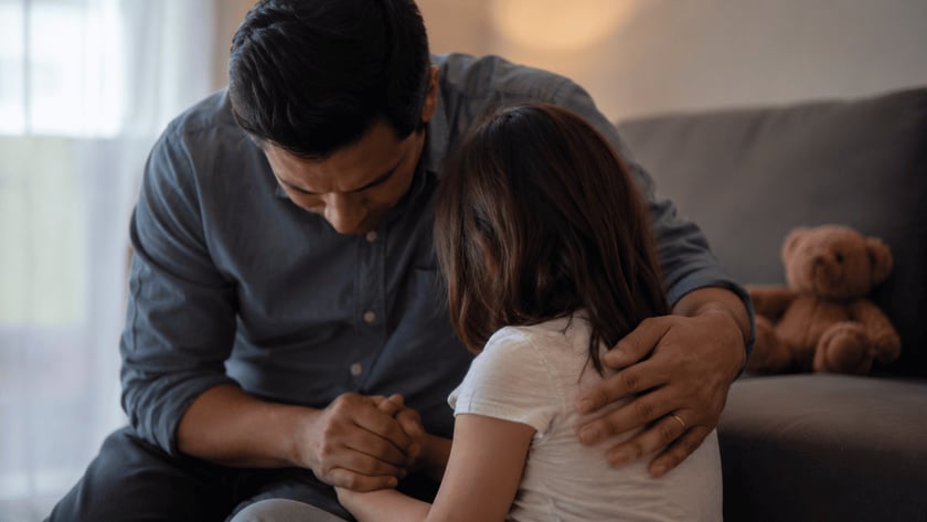 Homem de camisa azul-escura abraça uma menina de cabelo castanho, de mãos dadas, dentro de casa.