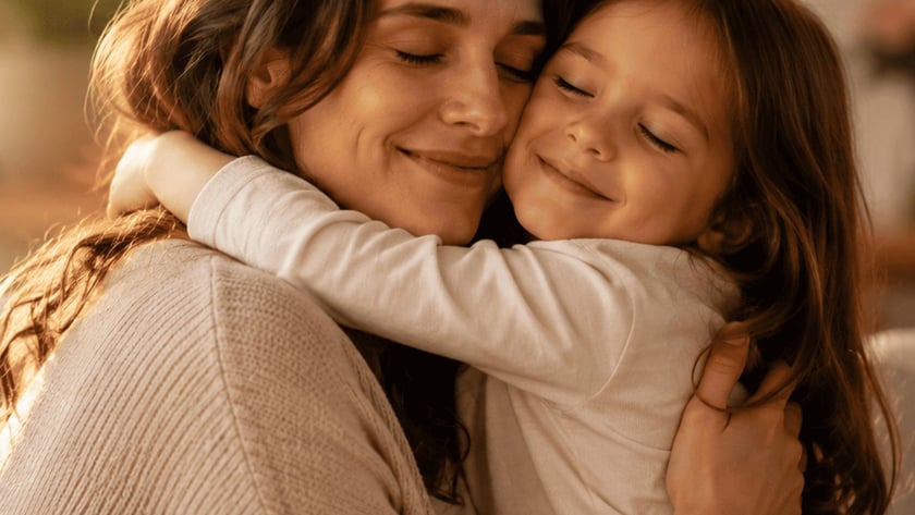 Mulher de cabelos castanhos abraça uma menina sorridente, ambas de olhos fechados, dentro de casa.