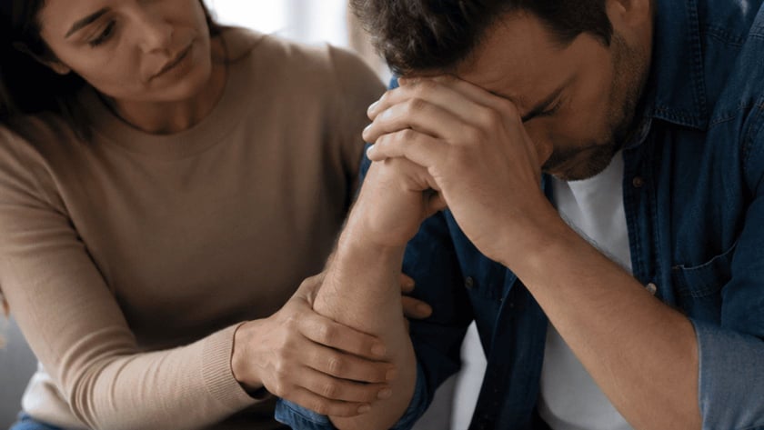 Mulher sentada em um sofá segura o braço de um homem de cabeça baixa.