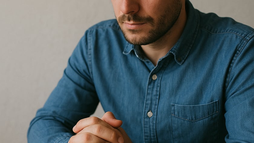 Homem de barba curta e camisa jeans, sentado à mesa, olha para as mãos entrelaçadas.