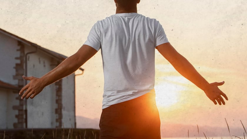 Homem de costas, com os braços abertos, olha o pôr do sol perto de uma casa.