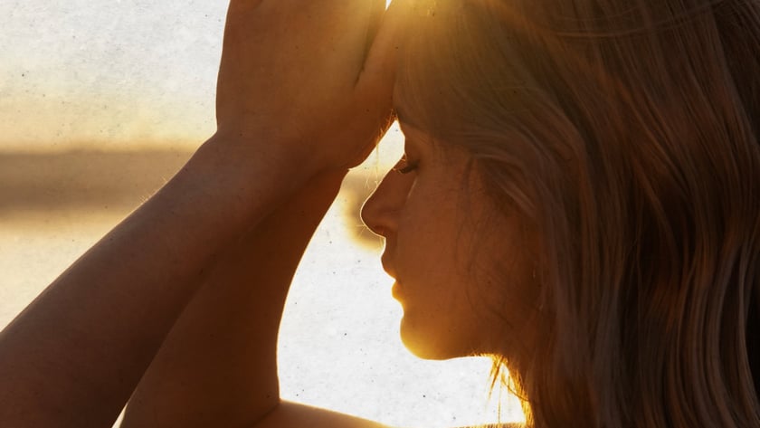 Mulher de perfil, de olhos fechados, com as mãos unidas acima da testa ao pôr do sol.