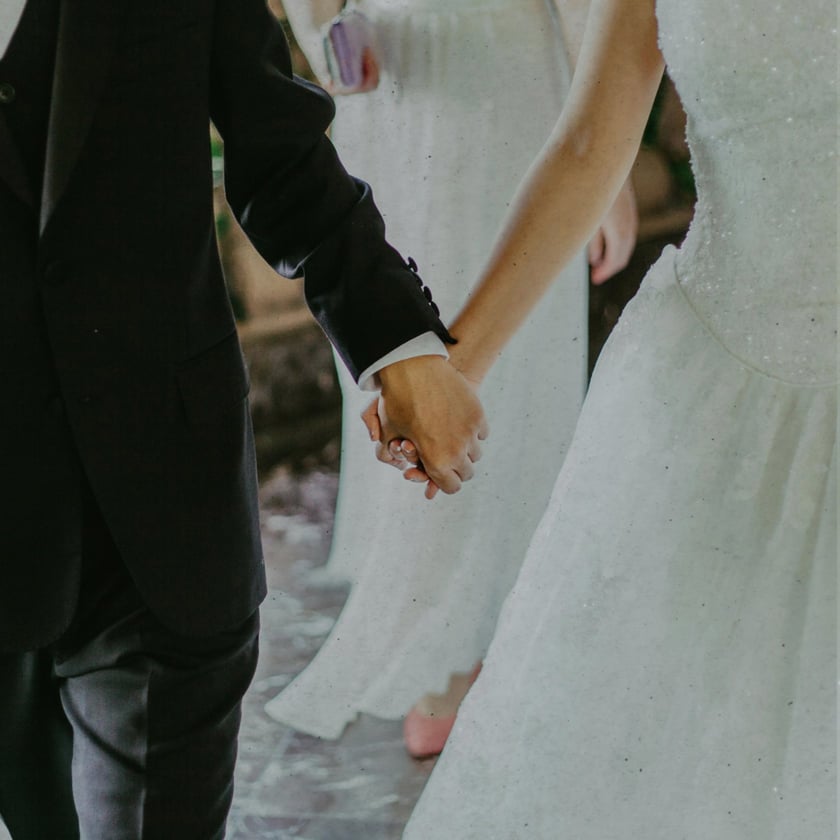 Noiva de vestido branco e noivo de terno escuro andam de mãos dadas.