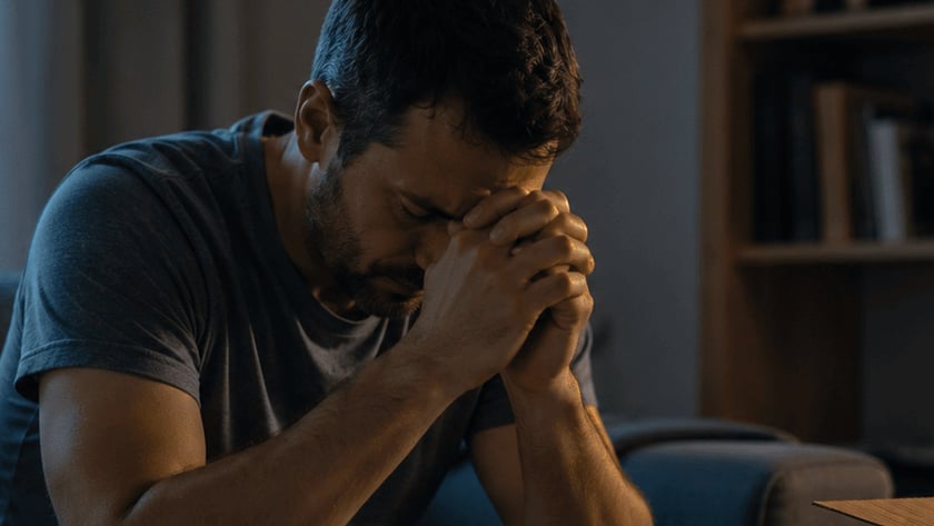 Homem sentado no sofá com as mãos unidas à testa e a cabeça baixa.