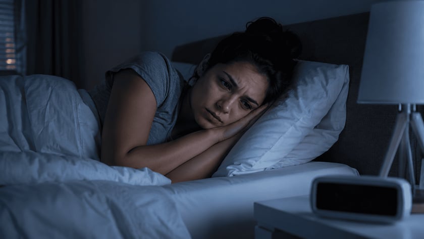 Mulher deitada na cama, de olhos abertos, olhando para o despertador na mesa de cabeceira.