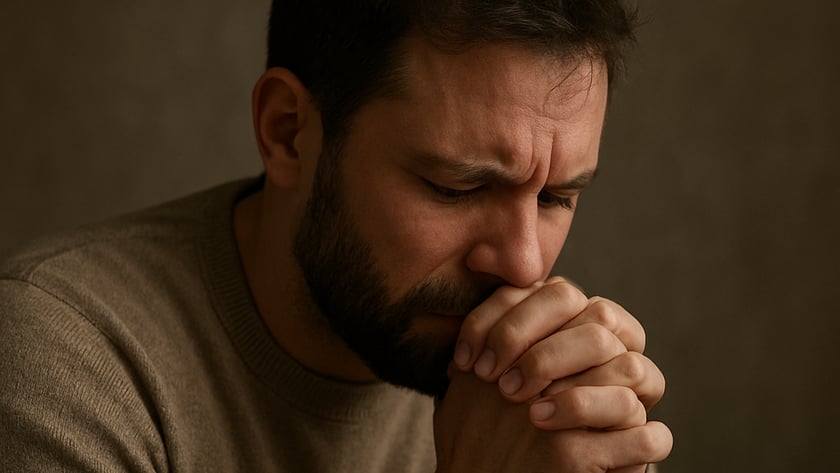 Homem de barba, com suéter marrom, de olhos fechados e mãos unidas junto ao rosto.