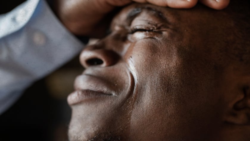 Homem de olhos fechados, com a mão na testa e lágrimas escorrendo pelo rosto.