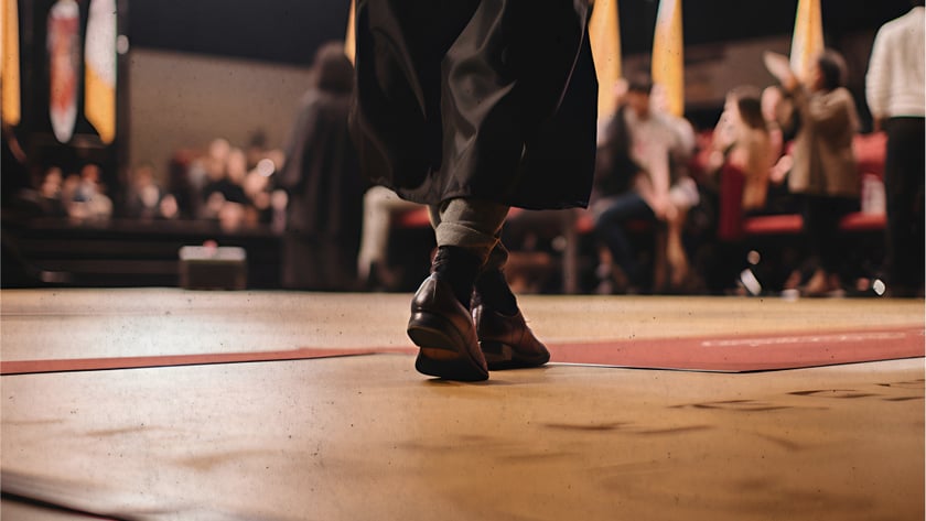 Pessoa de beca preta caminha em um palco de formatura diante da plateia desfocada.