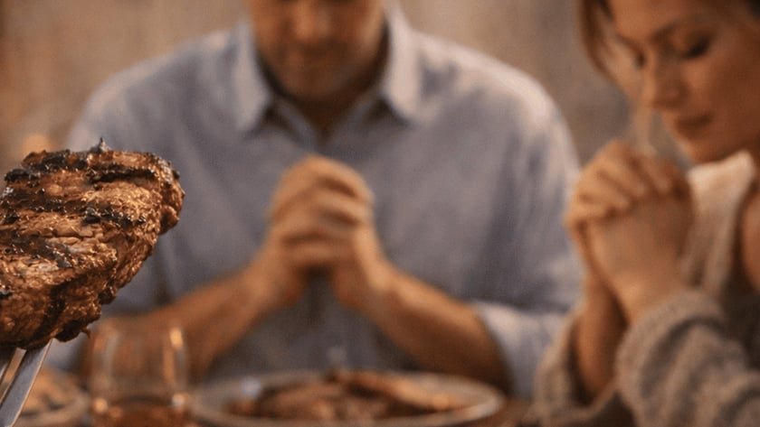 Homem de mãos postas, de cabeça baixa, à mesa com pratos de carne e um garfo em primeiro plano.