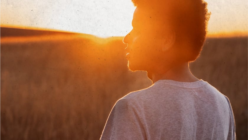Jovem de camiseta clara e cabelo cacheado olha de perfil para um campo de grama seca.