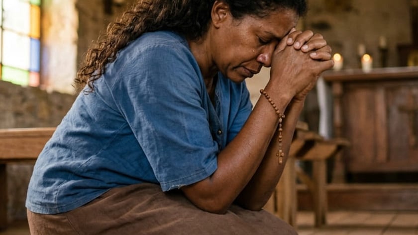 Mulher ajoelhada em uma igreja de pedra, com as mãos postas e um terço no pulso.