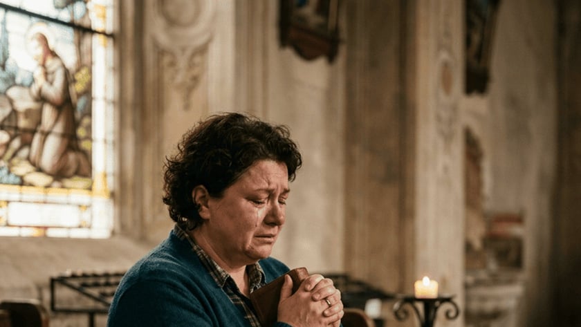 Mulher de cabelo curto e cardigã azul segura um livro junto ao peito, sentada em banco de igreja.