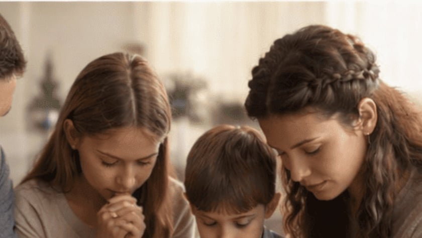 Mulher, menina e menino sentados juntos, de olhos fechados e mãos unidas, rezando.