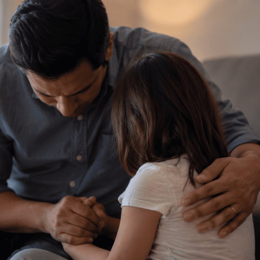 Homem de camisa azul-escura abraça uma menina de cabelo castanho, de mãos dadas, dentro de casa.