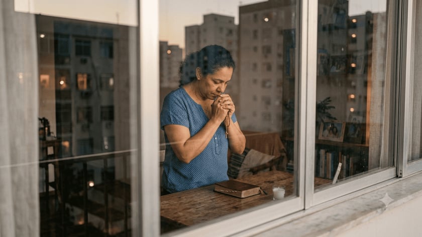 Mulher de blusa azul reza com um terço junto à janela, diante de uma Bíblia e uma vela.