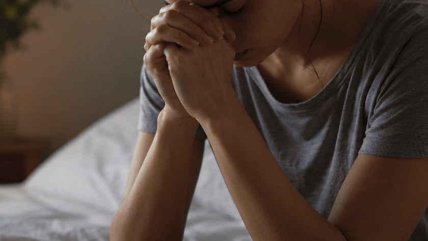 Mulher sentada na cama, com as mãos juntas diante do rosto, perto de um abajur aceso.