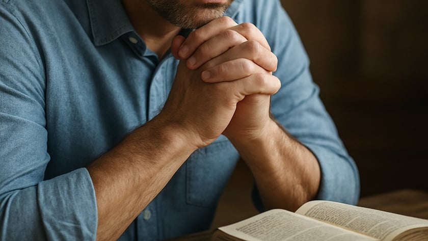 Homem de óculos e camisa azul, de olhos fechados, com as mãos unidas diante de um livro aberto.