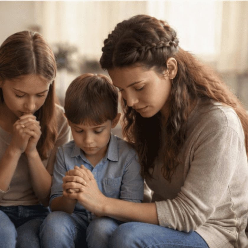 Mulher, menina e menino sentados juntos, de olhos fechados e mãos unidas, rezando.